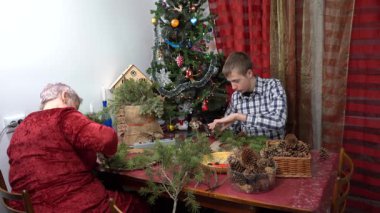Büyükannem ve torunum birlikte Noel çelengi yapıyorlar. Yaşlı kadın ve genç çocuk Noel ağacı dallarını ve konilerini yapıştırıyor. Aile içi kış aktiviteleri.