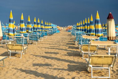 Güneş pansiyonları şemsiyelerle kumlu sahilde kürek çekiyor. Boş sahil beldesi. İtalya. Sahil şeridi şezlongunda güneş banyosu. Yazın mavi yağmurlu gökyüzü