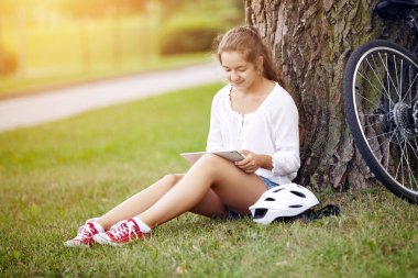 Güzel bir genç kız bir parkta çim üzerinde oturur ve bir tablet kullanır. Teknoloji Internet modern yaşam konsepti.
