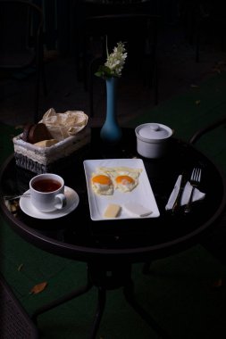 Kızarmış yumurtalı kahvaltı tabağı, siyah masada bir fincan çay ve ekmek.