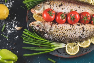 Bitkilerle dolu tam balık, domates ve limon dilimleri gri tabakta.