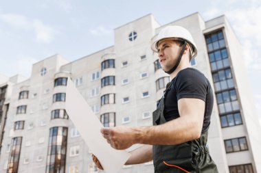 Miğferli bir müteahhit ve inşaat halindeki bir evin arka planında bir çizim var.