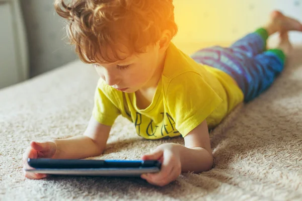 Pequeño niño lindo en una camiseta verde jugando juegos en una tableta y viendo dibujos animados. concepto de adicción — Foto de Stock