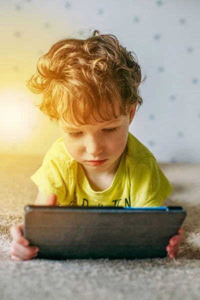 Маленький милый мальчик в зеленой футболке играет в игры на планшете и смотрит мультики. концепция зависимости — стоковое фото