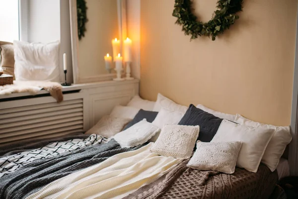 Bright cozy modern bedroom with holiday decoration. Bed with grey bedding set and knitted pillow on it, wooden rack, beautiful Christmas tree wreath on the wall and candle lights.