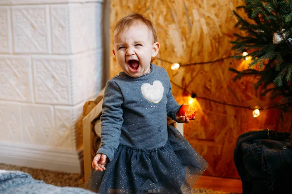 Imagem da menina doce e bonito em vestido cinza, retrato da bela criança sorridente de 1 ano de idade, criança. Menina feliz no interior decorado de Natal com tangerina na mão — Fotografia de Stock