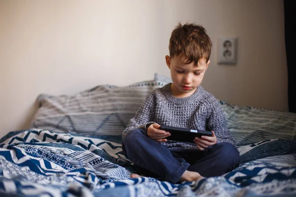 Netter kleiner Junge, der mit einem Smartphone auf dem Bett spielt. Konzept der Internetabhängigkeit — Stockfoto