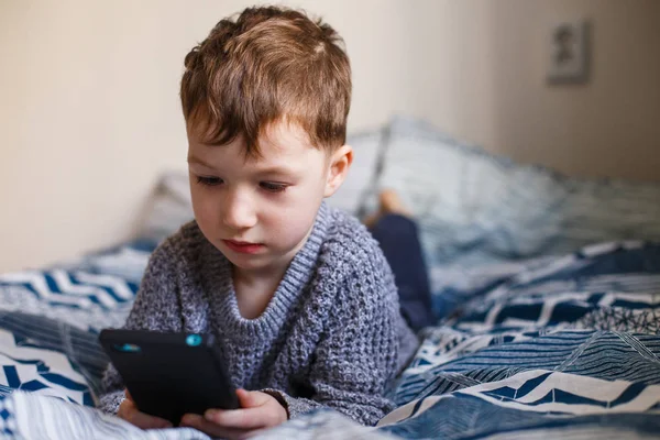 Netter kleiner Junge, der mit einem Smartphone auf dem Bett spielt. Konzept der Internetabhängigkeit — Stockfoto