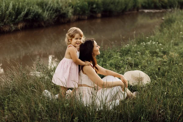 O familie fericită pe o pajişte de vară. Fetiță fetiță copil fiica îmbrățișare și sărut mama. Familia stând lângă râu — Fotografie, imagine de stoc