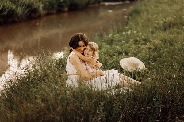 Yaz otlağında mutlu bir aile. Küçük kız bebek kız, sarılıyor ve annesini öpüyor. Aile nehir kenarında oturuyor.