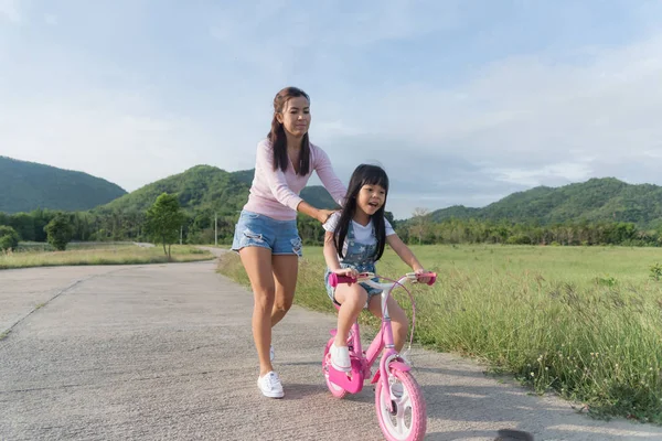 An encouraging mother helps her daughter learn how to steer her new bicycle. Wearing a pink helmet, the daughter is proud and happy, looking down while smiling.