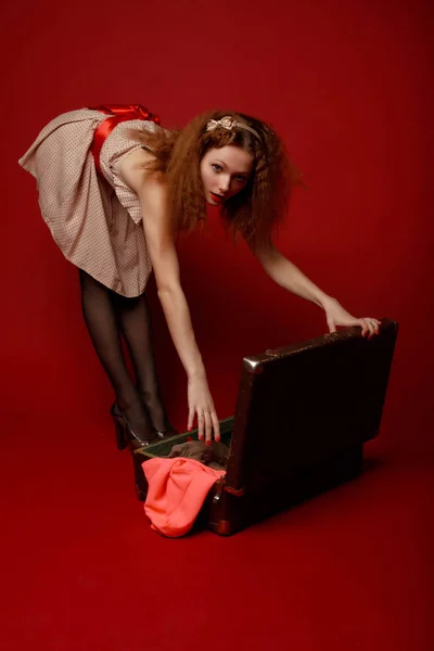 穿着短裙 红色腰带和高跟鞋的漂亮年轻女子在红色背景下把她掉落的衣服放在打开的行李箱上 引式全增长复古式肖像 — 图库照片
