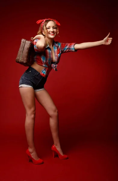 Pretty young woman in shorts,red high heels, red bandana and decolletage colorful shirt stands holding suitcase and asks for hitchhiking against red background. Full-growth retro-style pin-up portrait