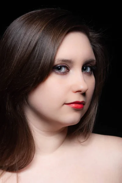 Beautiful gray-eyed brown-haired woman with red lips and bared shoulders against black background. Closeup studio portrait