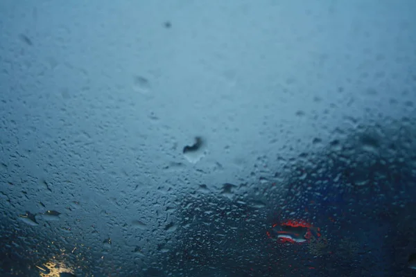 Cadute di pioggia sul parabrezza e luci sfocate delle auto. Concetto astratto di fondo — Foto Stock