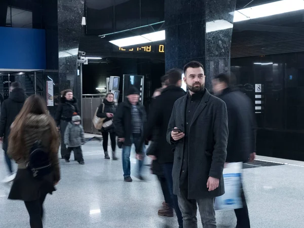 Handsome bearded man dressed in wool coat holds a smartphone and stands still in metro station within moving crowds of people — Stock Photo, Image