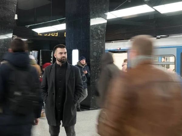 Handsome bearded man dressed in wool coat stands still in metro station within moving crowds of people — Stock Photo, Image