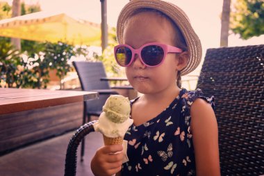 Hasır şapka ve pembe güneş gözlüğü takmış küçük bir kız kafede dondurma yiyor..