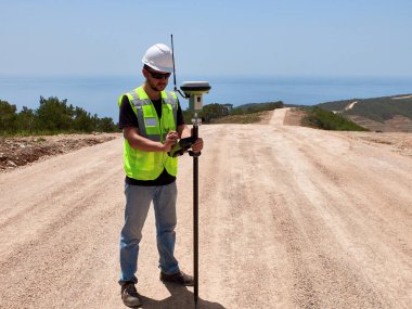 Geodetic engineer surveyor in white hard hat doing measurements with GNSS satellite receiver during road construction works. clipart