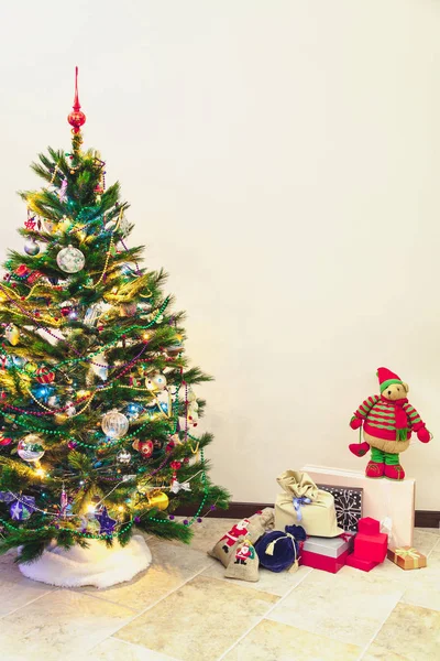 Attributes of the New Year holiday. A dressed tree with toys and garlands stands against a white wall. Next to the box with gifts. Above is a big soft to — Stock Image Attributes New Year Holiday Dressed Tree Toys Garlands Stands White Royalty Free Stock Photos