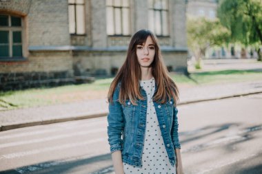 Uzun saçlı, kot ceketli sevimli genç bir öğrenci sonbahar öğleden sonraları kampüste yürüyor.