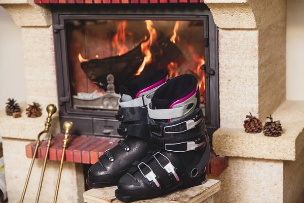 在壁炉前烘干鞋子 两只滑雪靴站在一个燃烧 Fireplac 前的凳子上 — 图库照片