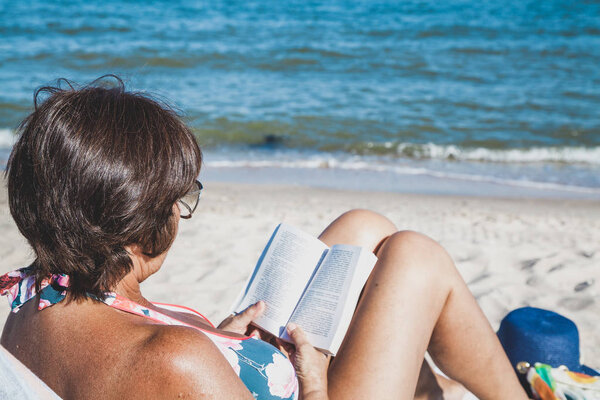 Elderly woman in a swimsuit sunbathing and reading a book on summer sea beach