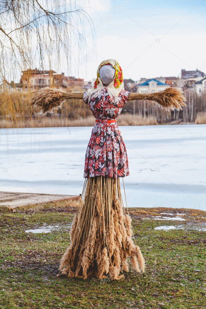 Slavic holiday of the end of winter. A large Shrovetide doll made of straw in a woman's dress and scarf stands on the river ban