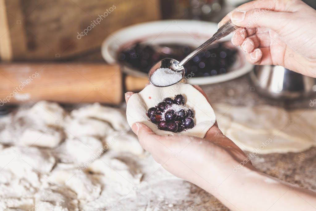 woman hand pours sugar into a raw dumpling with a stuffing of black round berries. On the table lie raw dumplings, sieve and rolling pi