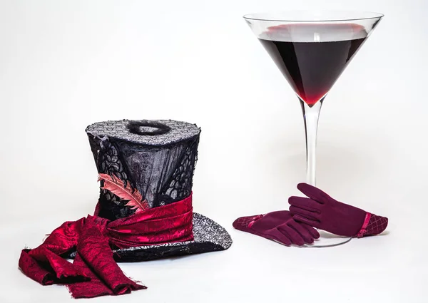 Magic still life. A big cylinder hat and a huge glass goblet with red wine on a white background. Near red women\'s glove