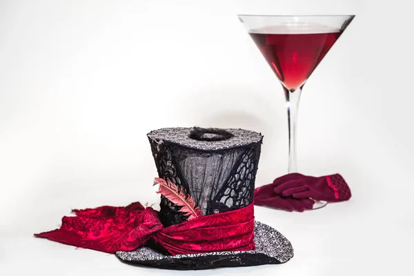 Magic still life. A big cylinder hat and a huge glass goblet with red wine on a white background. Near red women's glove
