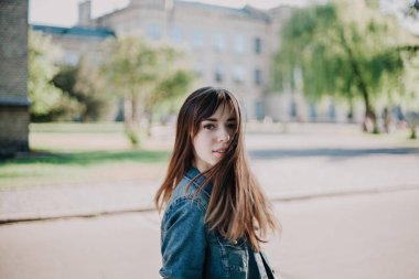 Uzun saçlı, kot ceketli sevimli genç bir öğrenci sonbahar öğleden sonraları kampüste yürüyor.