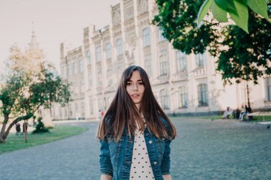 Uzun saçlı, kot ceketli sevimli genç bir öğrenci sonbahar öğleden sonraları kampüste yürüyor.