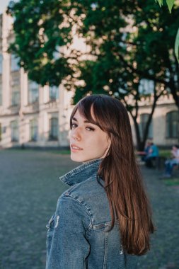 Uzun saçlı, kot ceketli sevimli genç bir öğrenci sonbahar öğleden sonraları kampüste yürüyor.