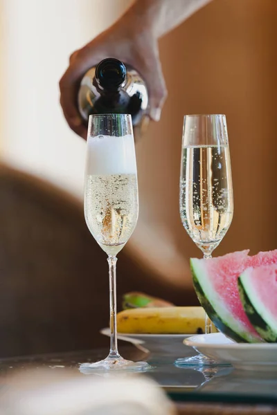Romantic plot with champagne. The hand pours a sparkling wine with a thin stream into tall, elegant glasses. Next to fruit and watermelo