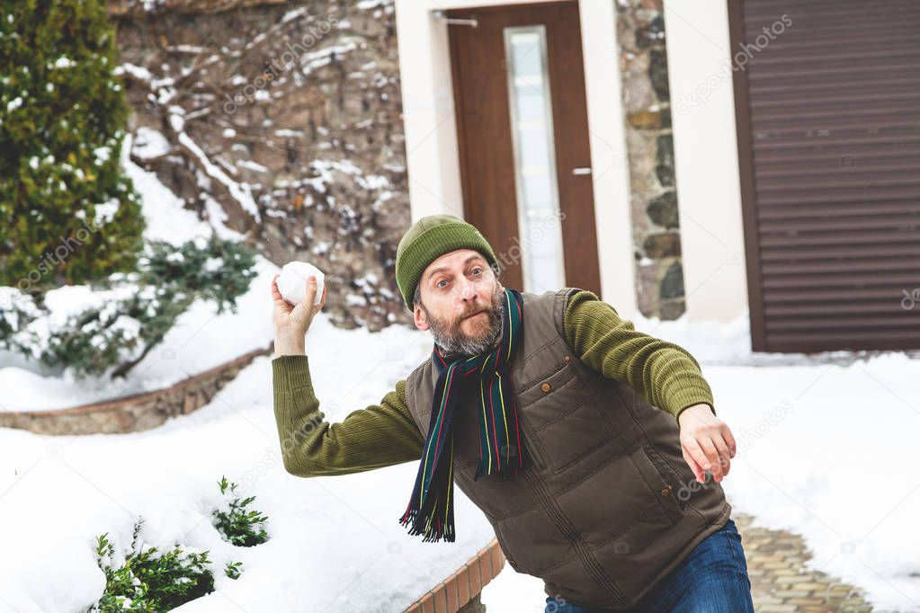 Snow games on the air. A man with a beard in a warm waistcoat and hat throws snowballs in the courtyard of a private house in a frosty da