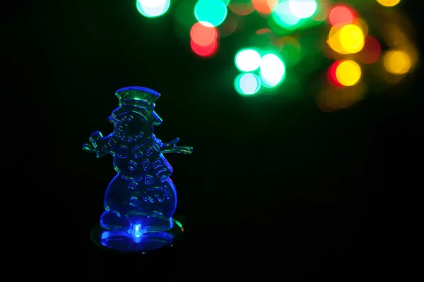 Transparent night light in form of snowman is lit in blue on black background. Bokeh of colorful lights in background