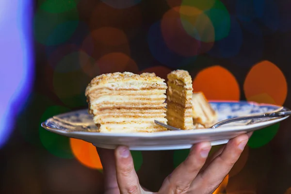 Appetizing piece of cake lies on a plate close-up. In the background blurred bright lights of Christmas garland