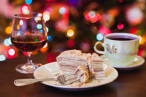 Christmas dessert still life. Appetizing piece of cake lies on a plate close-up. Near a glass of brandy cup of te
