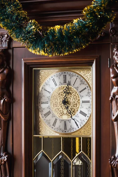 Vintage pendulum clock in a brown wooden carved case is decorated with Christmas garland close u