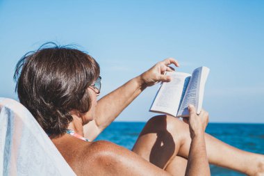 Yaşlı kadın bir mayo güneşlenme ve yaz deniz plaj üzerine bir kitap okuma