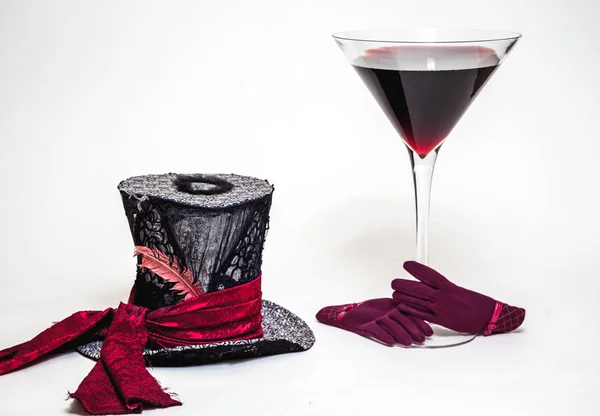 Magic still life. A big cylinder hat and a huge glass goblet with red wine on a white background. Near red women\'s glove