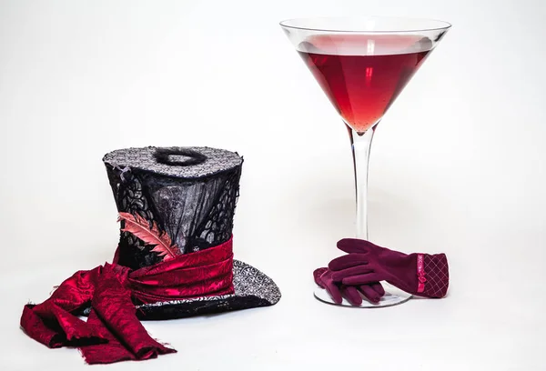 Magic still life. A big cylinder hat and a huge glass goblet with red wine on a white background. Near red women\'s glove