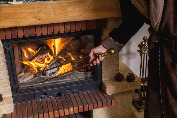 man in a warm robe holding a large brass fireplace tongs. In a fireplace fire burns brigh
