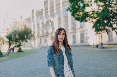 Uzun saçlı, kot ceketli sevimli genç bir öğrenci sonbahar öğleden sonraları kampüste yürüyor.