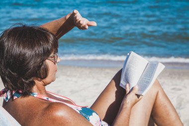 Yaşlı kadın bir mayo güneşlenme ve yaz deniz plaj üzerine bir kitap okuma