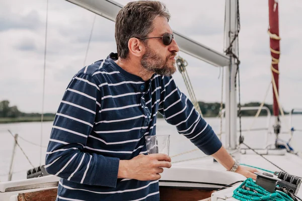 Человек с бородой в очках пьет виски на яхте — стоковое фото