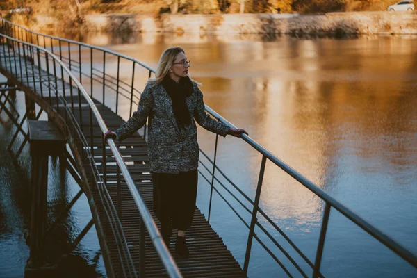Κομψή ξανθιά στέκεται σε στενή γέφυρα πάνω από τη λίμνη του φθινοπώρου — Φωτογραφία Αρχείου