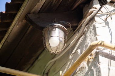 Beyaz bir duvarın çatısının altında yarı dairesel bir gölgesi olan lamba. Yakınlarda elektrik kabloları ve gaz borusu var.