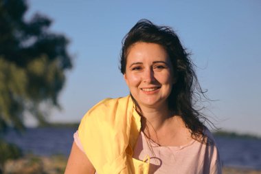 Geniş bir nehrin kumlu sahilinde uzun kızıl saçlı ve parlak sarı ceketli güzel bir kızın portresi.